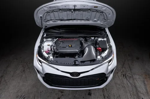 Car engine compartment with hood open, revealing various components including a carbon fiber air intake system, set in a dimly lit garage with concrete floor.