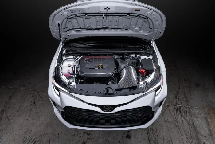 Car engine compartment with hood open, revealing various components including a carbon fiber air intake system, set in a dimly lit garage with concrete floor.