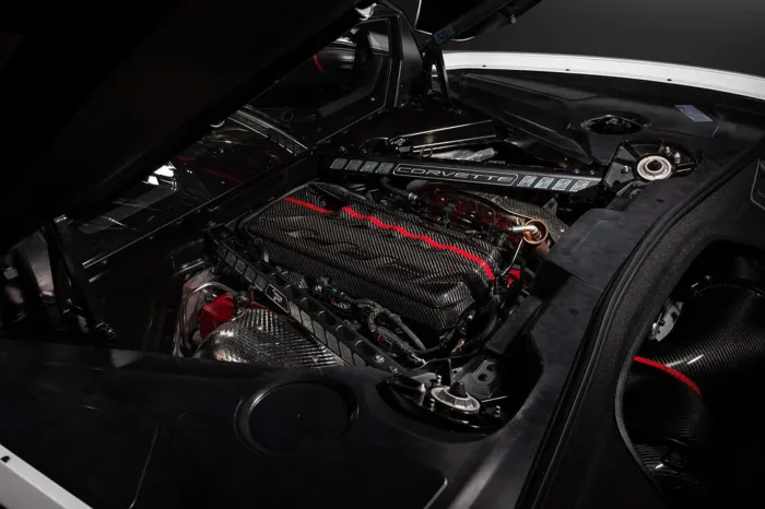 Car engine with a carbon fiber cover sits inside a sleek engine bay. The word "Corvette" is visible on a component above the engine, against a backdrop of black interior and metallic accents.
