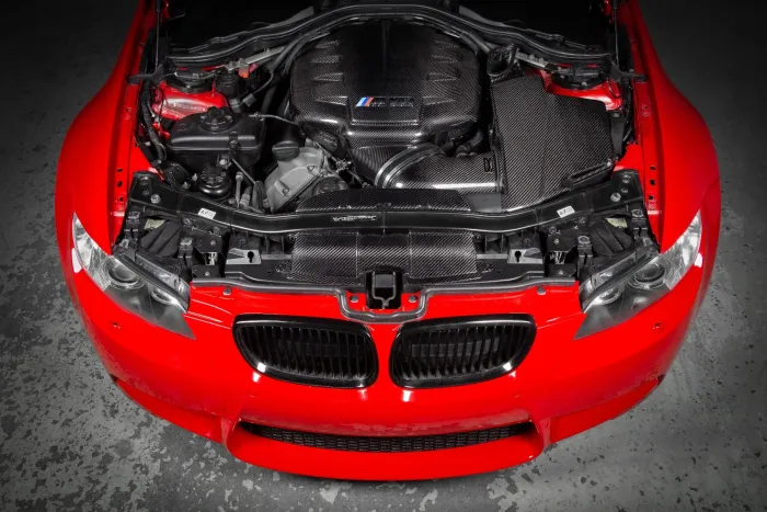Red car hood open, exposing a complex engine with carbon fiber components, in a garage setting with a concrete floor. The engine has a logo with three colored stripes.