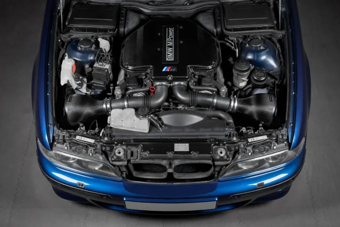 Engine displayed top-down in a blue car's hood, marked "BMW M Power." Nearby components include air filters, coolant reservoir, hoses, and electric elements, all in a garage setting.