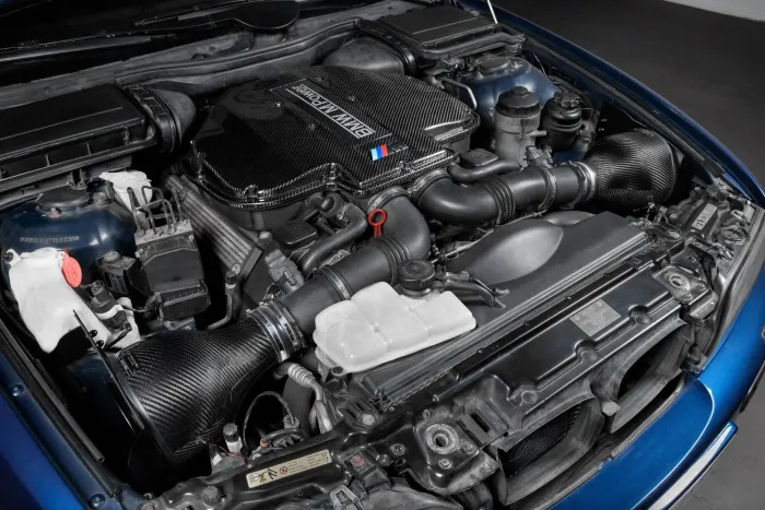 A high-performance BMW engine with a carbon fiber cover and various components visible, situated inside a car’s engine bay. "BMW POWER" is written on the engine cover.