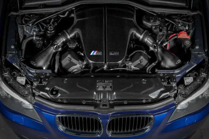 The engine bay of a blue BMW features a carbon fiber engine cover with the "M" logo and "V10" text, surrounded by mechanical components and hoses.