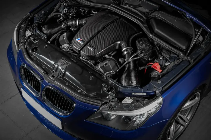A car engine with carbon fiber coverings, equipped with M Power components, sits inside the engine bay of a blue vehicle in a garage.