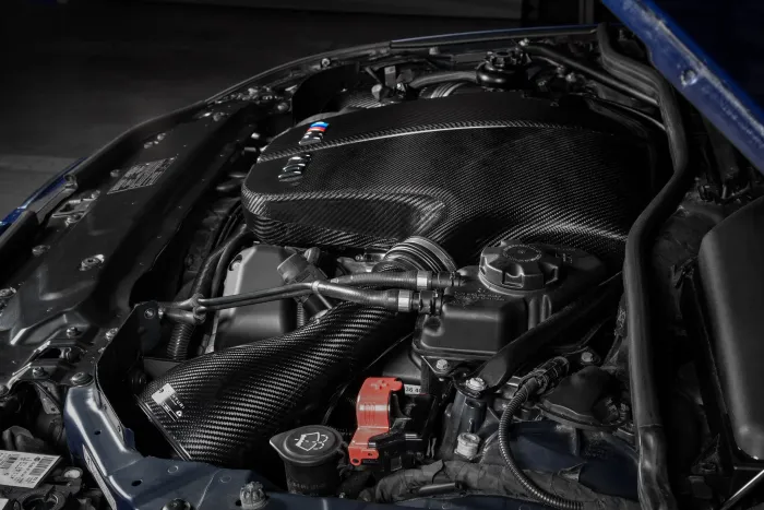 A high-performance car engine, featuring sleek, carbon-fiber components and various cables, is housed within the engine bay of a blue vehicle, with a dark surrounding environment.