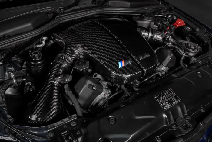 Carbon fiber engine cover and parts in a car's engine bay featuring a "BMW" logo with stripes (blue, purple, and red), surrounded by various cables, hoses, and components.