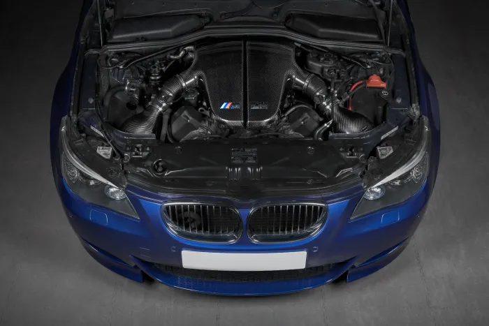A blue car with its hood open, revealing a detailed, high-performance engine featuring carbon fiber components. The car is positioned in a dimly lit garage.
