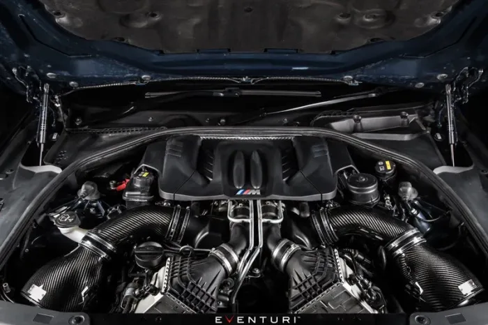 High-performance car engine with twin air intake pipes sits within an open hood, featuring the "M" logo and surrounded by metallic components. Text at the bottom reads: "EVENTURI".