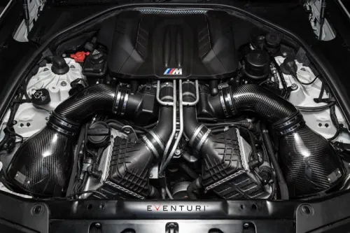 A high-performance BMW engine bay featuring carbon fiber intake components and the "M" logo, surrounded by various engine parts and housed within the car's chassis. The text "EVENTURI" is visible at the bottom.