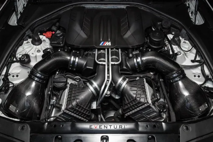 A high-performance BMW engine bay featuring carbon fiber intake components and the "M" logo, surrounded by various engine parts and housed within the car's chassis. The text "EVENTURI" is visible at the bottom.