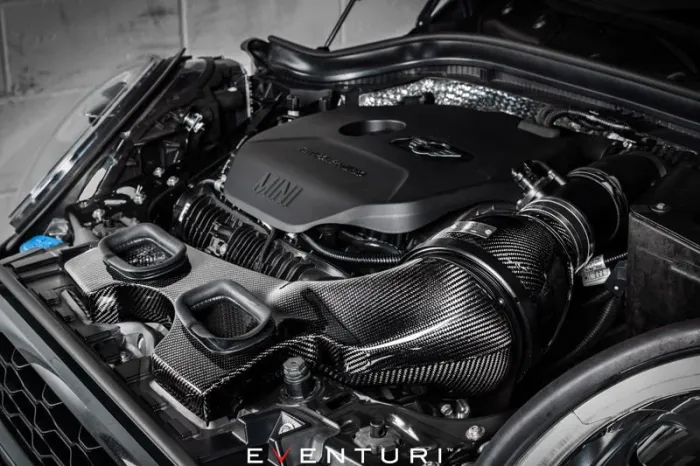 Engine covered in black casing labeled "MINI" and "Twin Power Turbo," integrated with carbon fiber intakes and piping by Eventuri, displayed in a car's open hood against a garage backdrop.