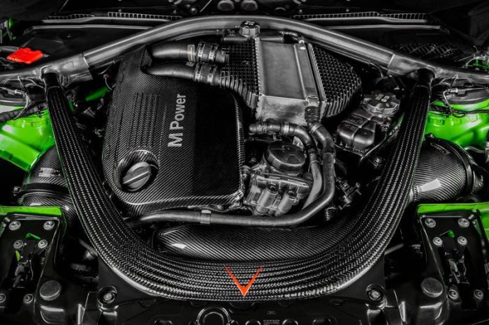 A high-performance engine with "M Power" branding is surrounded by various hoses and components, all encased in a sleek, black carbon fiber cover with bright green accents in the engine bay.