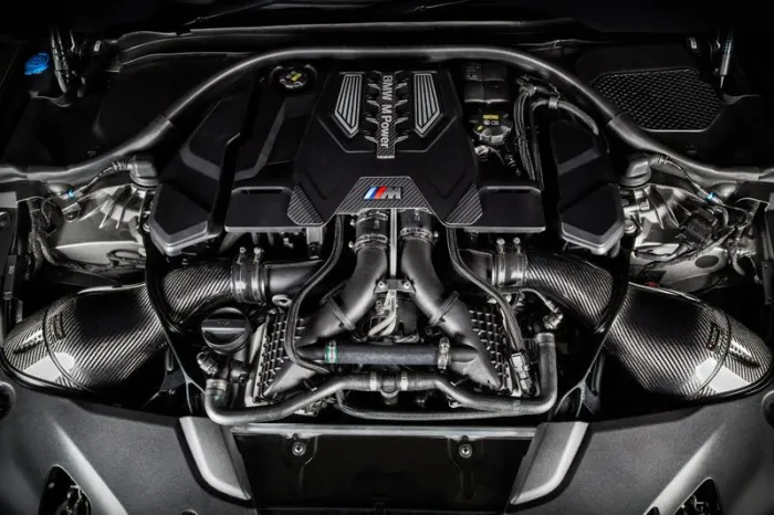 A high-performance engine with multiple carbon fiber components and visible intake manifolds; text reads "BMW M Power". The engine is mounted inside a car's engine bay.