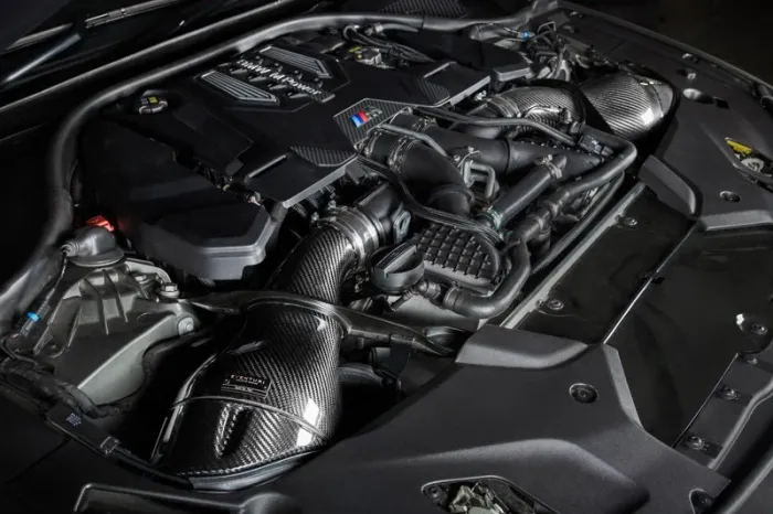 A high-performance engine with carbon fiber components is installed in a car's engine bay, displaying intricate black and silver parts with precision engineering details.