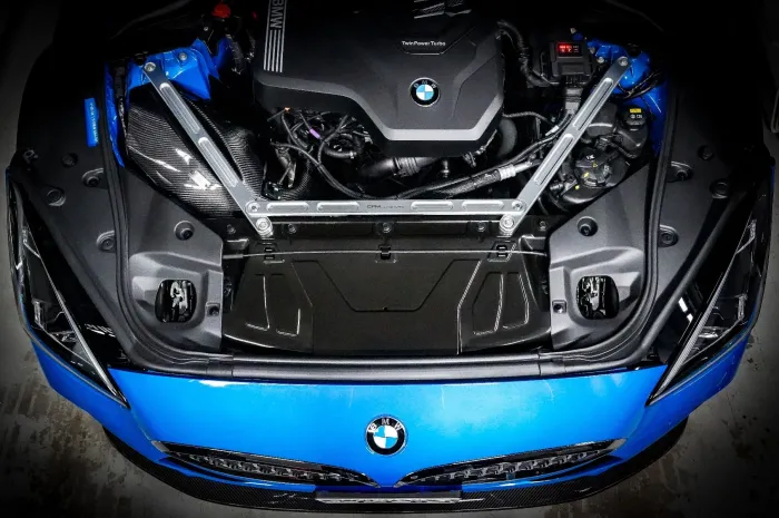 A BMW engine with "TwinPower Turbo" text displayed, mounted in the front of a blue car, secured with metallic struts and surrounding components, viewed from above in a garage setting.