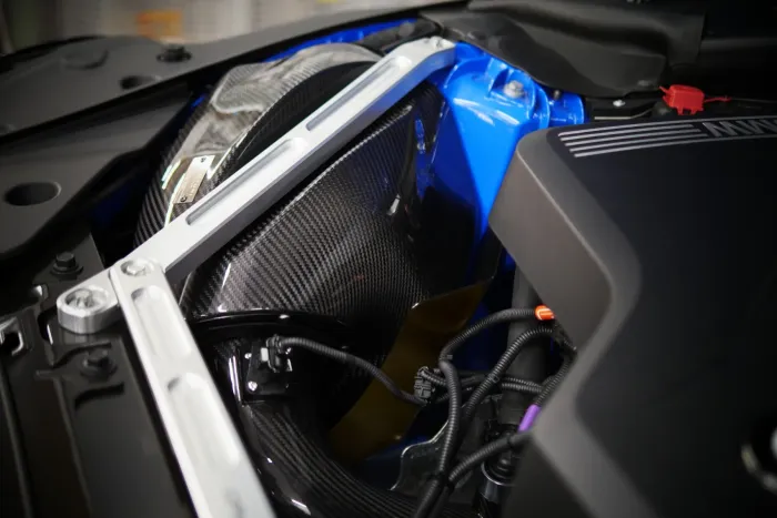 A section of a car engine bay featuring carbon fiber parts, blue and black components, and metal brackets with various wires connected, highlighting the complex mechanical structure.