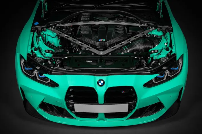 A teal BMW car with its hood open, revealing a detailed view of the engine bay, featuring a carbon fiber engine cover and structural support braces, against a dark background.