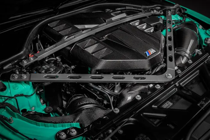 A high-performance car engine with carbon fiber components, marked with a blue, violet, and red "M" badge, encased in a green chassis, and supported by metallic struts.