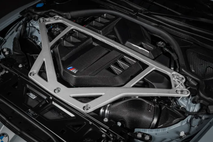A silver strut bar reinforces a black, carbon fiber engine cover marked with the M logo, within a modern vehicle's engine bay featuring various components and connections.