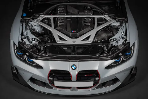 A BMW car with its hood open displays a powerful engine enclosed in a carbon-fiber cover, indicated by the BMW logo and 'M' badge, with some structural components visible.