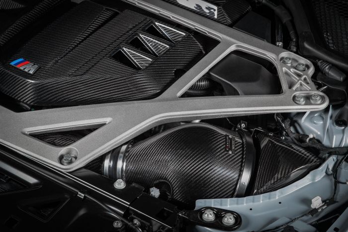 Carbon fiber engine cover with an "M" logo under a metallic strut brace; part of a high-performance vehicle's engine bay with visible wiring and components.