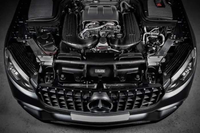 A sleek car engine with visible metallic components and carbon fiber coverings, housed inside a car with a prominent Mercedes-Benz grille in a garage setting.