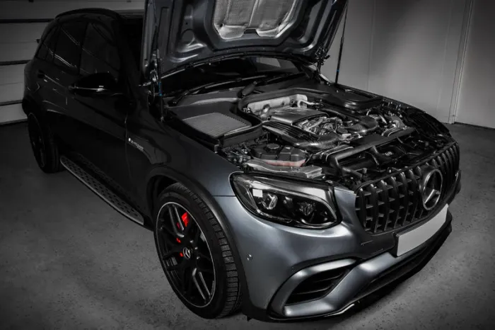 A luxury car with its hood open reveals a detailed engine compartment, parked inside a clean, well-lit garage with white walls.