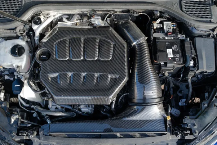 Carbon fiber engine cover and air intake system within a car engine bay, surrounded by various mechanical components, wiring, and a battery with visible labels and connectors.