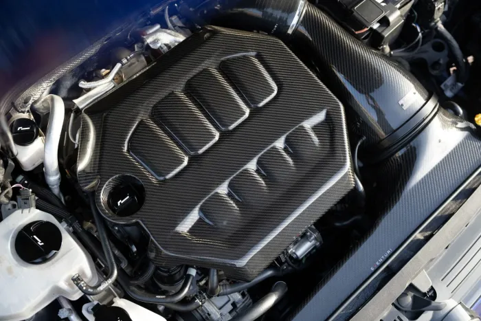 A car engine features a prominent carbon fiber cover with vent openings. The surrounding environment includes various tubes and components. The text "EVENTURI" is visible on a carbon fiber part.