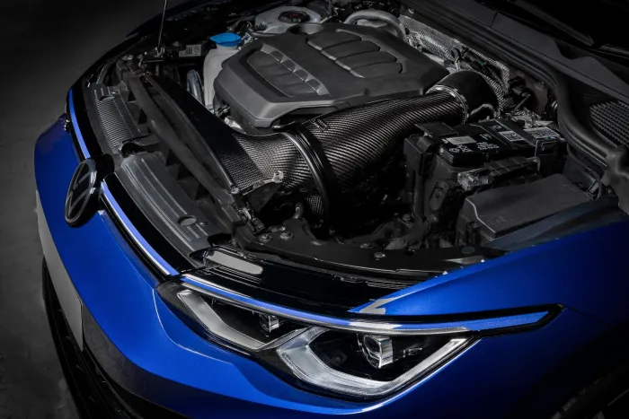 Car engine with a carbon fiber air intake system is installed in the front compartment of a blue vehicle with sleek headlight design, in a dimly lit garage.