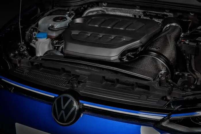 Volkswagen engine is encased in dark plastic with visible fluid reservoirs and a prominent carbon fiber intake in an open hood of a blue vehicle. The VW logo is centered below.