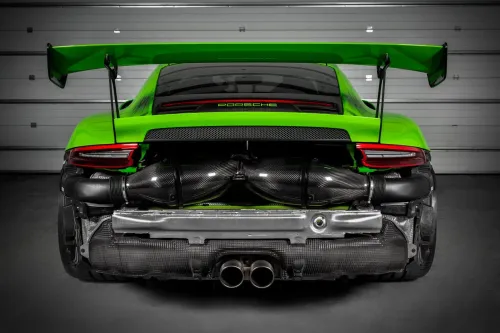 Green Porsche sports car with an elevated rear wing, large exhaust pipes, and visible engine components, parked in a garage with a closed door. The word "PORSCHE" is displayed on the back.