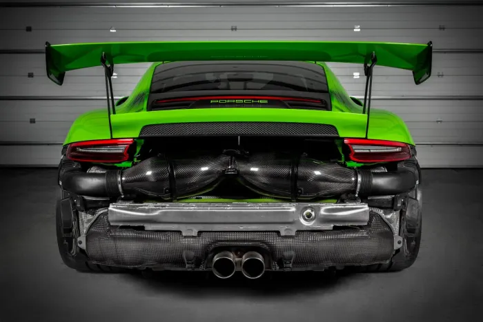 Green Porsche sports car with an elevated rear wing, large exhaust pipes, and visible engine components, parked in a garage with a closed door. The word "PORSCHE" is displayed on the back.