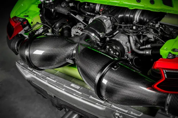 Carbon fiber air intake in a car engine bay with green panels and various mechanical components, including belts, hoses, and an alternator, in an indoor setting. Text on the intake reads "EVENTURI".
