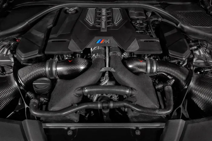 The BMW engine is situated in a car's engine bay, featuring "BMW M Power" text on the cover and surrounded by carbon fiber components and various hoses.