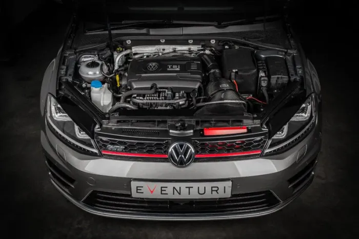 Car's engine bay with hood open. The Volkswagen engine is branded "TSI." The car has a grey exterior and a front license plate that reads "EVENTURI" in a dimly lit garage setting.