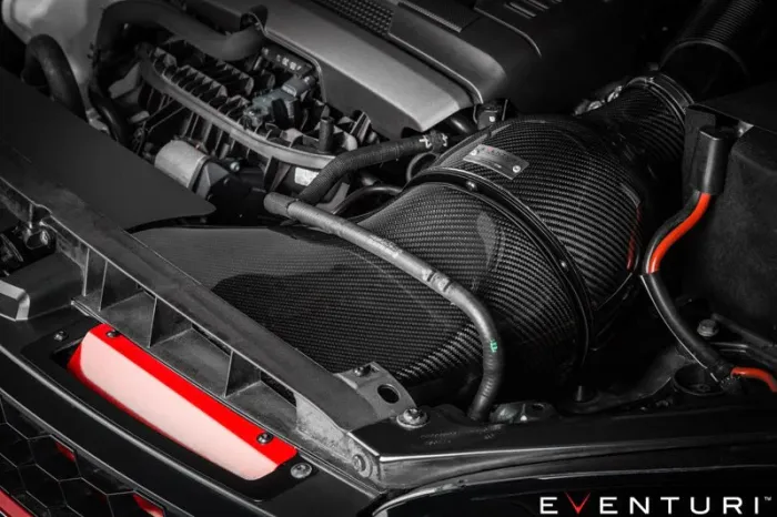 Carbon fiber air intake system situated in a car engine compartment with surrounding metallic components and tubes. Text "EVENTURI" is visible at the bottom right corner.