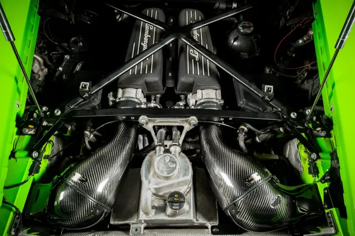 Lamborghini-branded V12 engine block with twin air intakes and carbon-fiber components sits within a bright green engine bay, emphasizing the high-performance mechanics and sporty design of the vehicle.