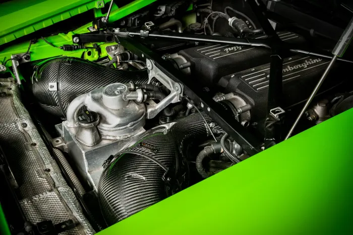 Engine compartment with "Lamborghini" on dual black air intakes, metallic components, and braided carbon-fiber pipes, surrounded by green-painted framework.