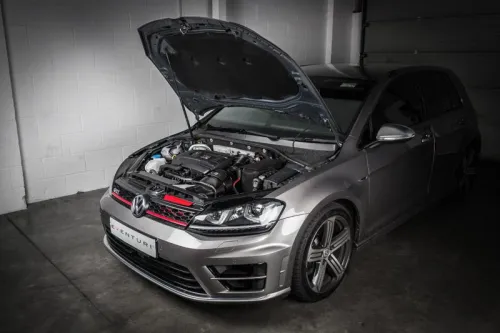 A gray Volkswagen car with the hood open reveals its engine components. The car sits in a dimly lit garage with white brick walls. License plate reads "EVENTURI".