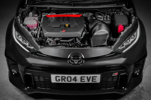 Car engine bay containing a visible engine with red and black components, and a front license plate reading "GR04 EVE", set within a black Toyota vehicle in an indoor setting.