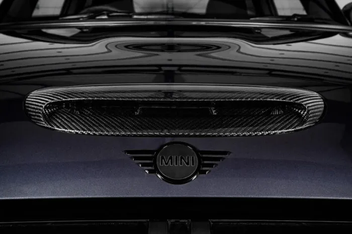 A carbon fiber hood scoop and "MINI" emblem on a sleek, dark-colored surface, reflecting ceiling patterns, likely part of a Mini Cooper car's exterior.