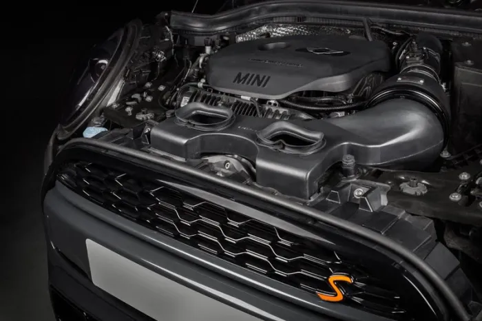 Car engine labeled "MINI," featuring a prominent airflow system and various mechanical components, situated within the open hood of a vehicle with a visible front grille and an orange "S" badge.