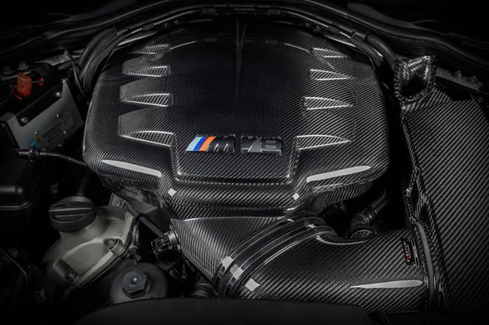 A carbon fiber engine cover labeled "M Power" sits at the center of a car engine bay, with various components and wires surrounding it, signifying a high-performance vehicle.