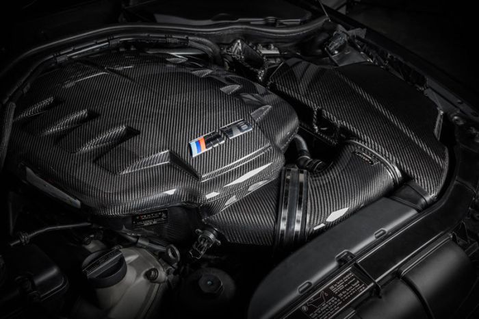 Carbon fiber engine cover with a BMW M Power logo on a high-performance car engine in a dark, industrial garage setting.