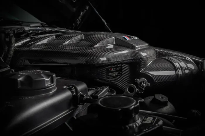 Carbon fiber engine cover with Eventuri branding sits in a dimly lit car engine compartment with various mechanical components surrounding it. Text on the cover reads "Eventuri, B58 Carbon Plenum."