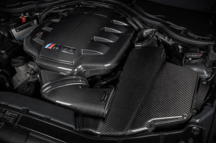 Carbon fiber engine cover labelled "M5" with colored stripes in a car engine bay; visible surrounding components include various cables and other engine parts.