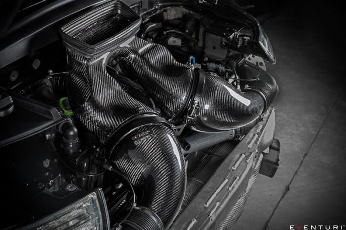 Carbon fiber air intake installed inside a car engine bay, featuring smooth, sculpted designs and labeled "Eventuri." The car’s front bumper and adjacent engine components are visible in a dimly lit garage setting.