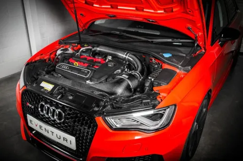 A bright orange Audi car with an open hood revealing the engine. The front grille features an "EVENTURI" license plate. The vehicle is positioned in a garage with gray concrete walls.