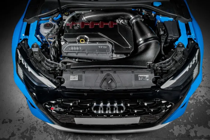 A car engine with "Audi TFSI" branding and red accents is installed under the open hood of a blue vehicle, with surrounding components and cables visible.